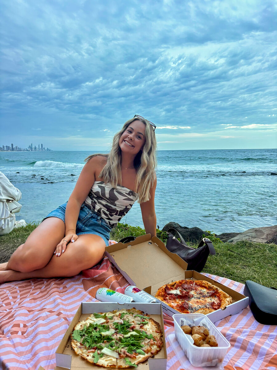 Gem sitting on a picnic rug at the beach with pizza